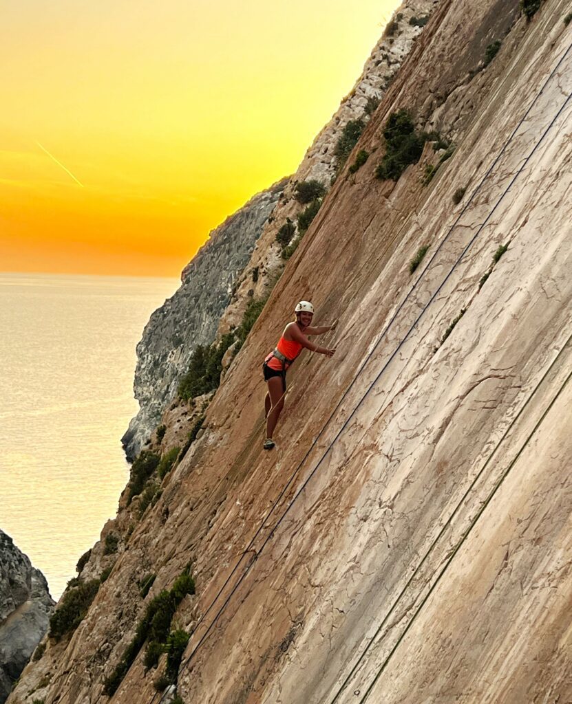 Woman climbing on a limestone wall with an orange sunset glowing over the sea