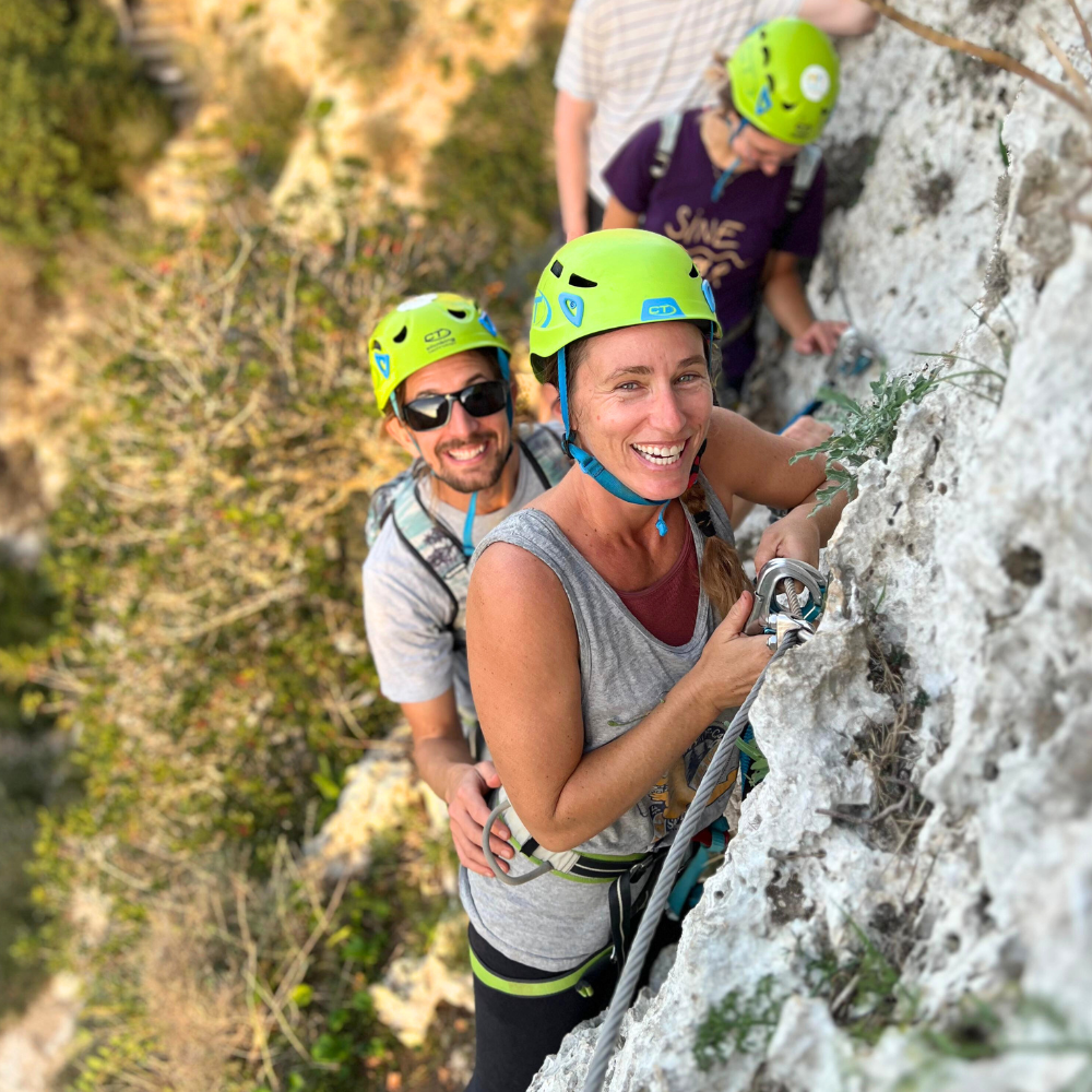 Couple smiling while climbing the Via Ferrata in Malta, attached to the safety cable on a cliffside route.