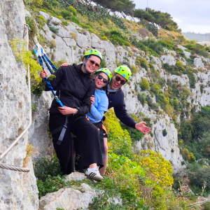 Family climbing the Via Ferrata in Malta, all clipped to the safety cable on the rock face.